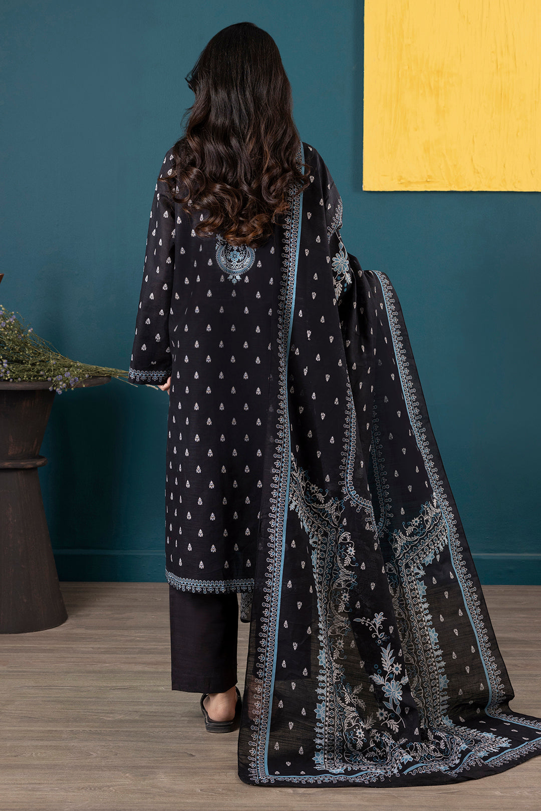 Woman wearing a black embroidered traditional outfit with a matching dupatta against a blue and yellow wall.