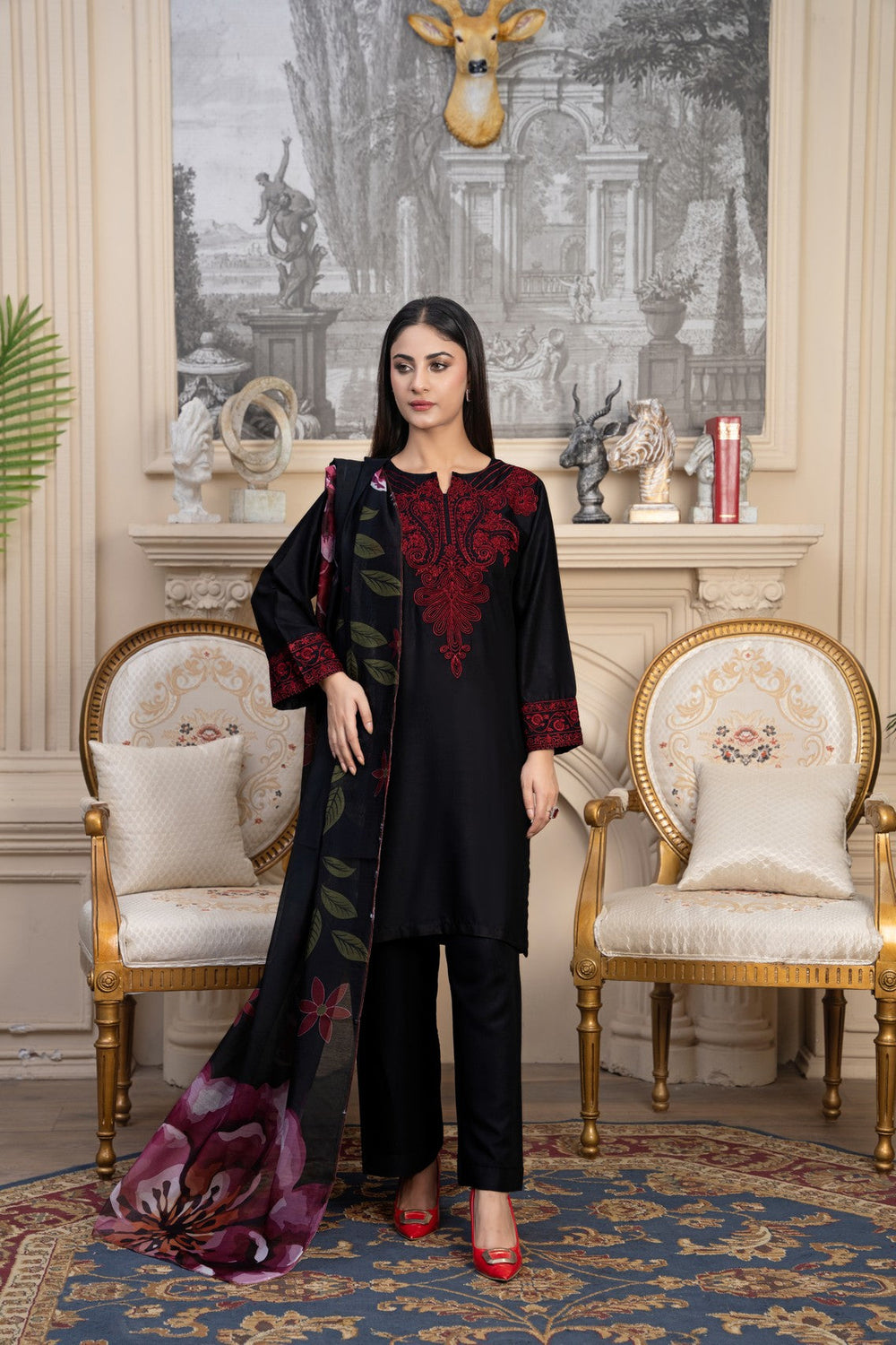 Woman in a black outfit with red embroidery standing in an elegant room with decorative chairs and artwork.