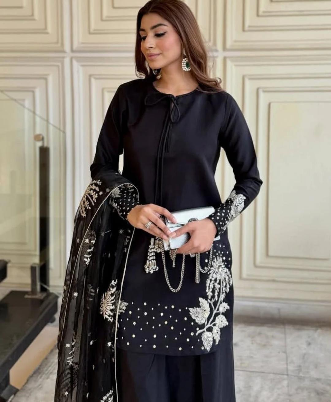 Woman in a black outfit with white embroidery standing in an elegant interior setting.