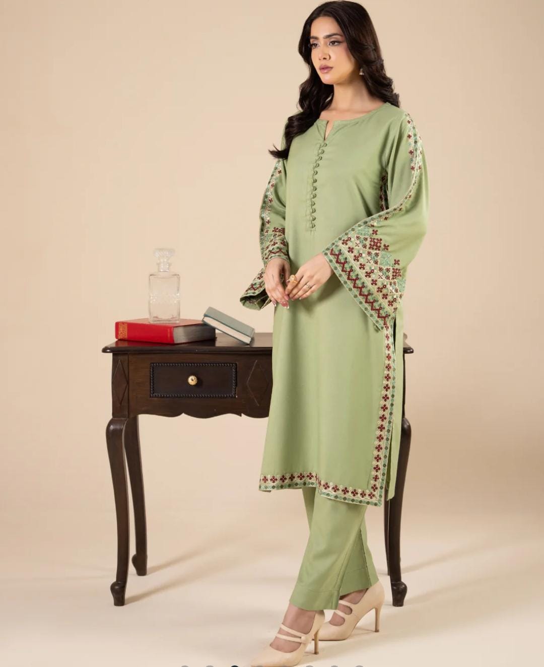 Woman wearing a light green traditional outfit with embroidery, standing in front of a wooden table.