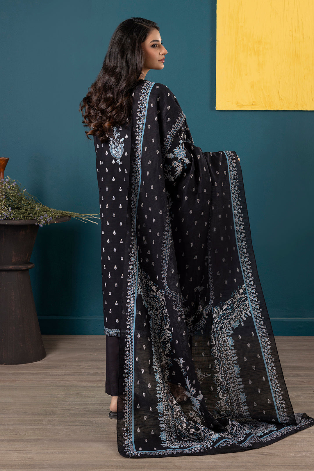 Woman wearing a black embroidered traditional outfit with a blue wall and plant in the background