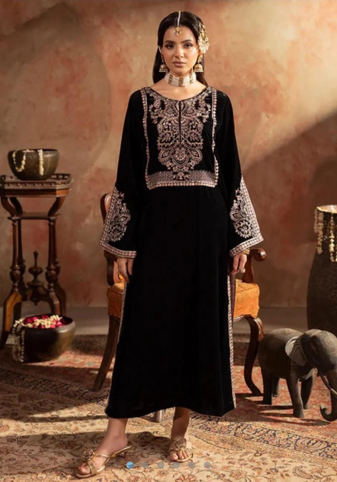 Woman in a black embroidered dress standing in an ornate room with a decorative background.