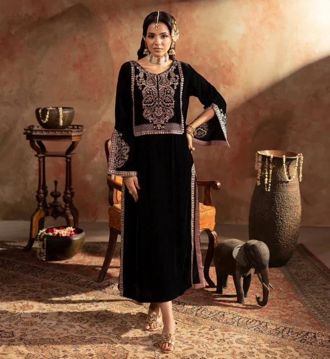 Woman in traditional black embroidered dress standing in a rustic room with decorative elements.