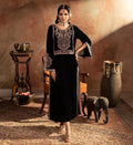 Woman in traditional black embroidered dress standing in a rustic room with decorative elements.