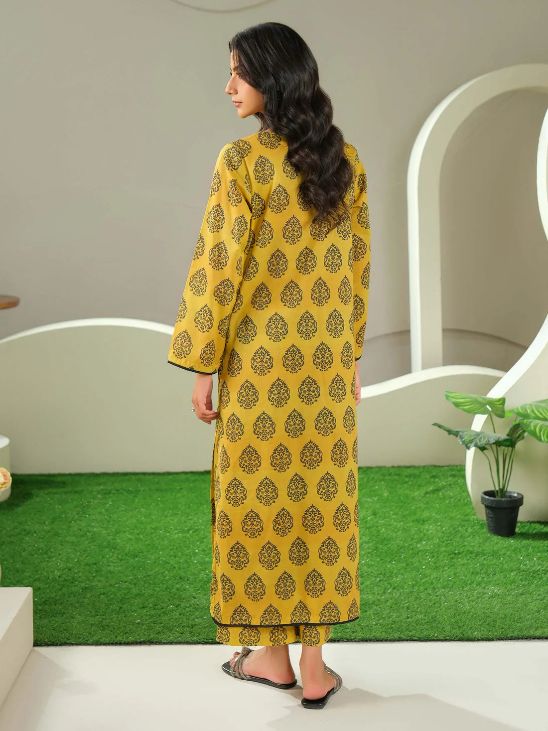 Woman wearing a yellow patterned kurta standing in a modern interior setting with green grass effect on the floor.