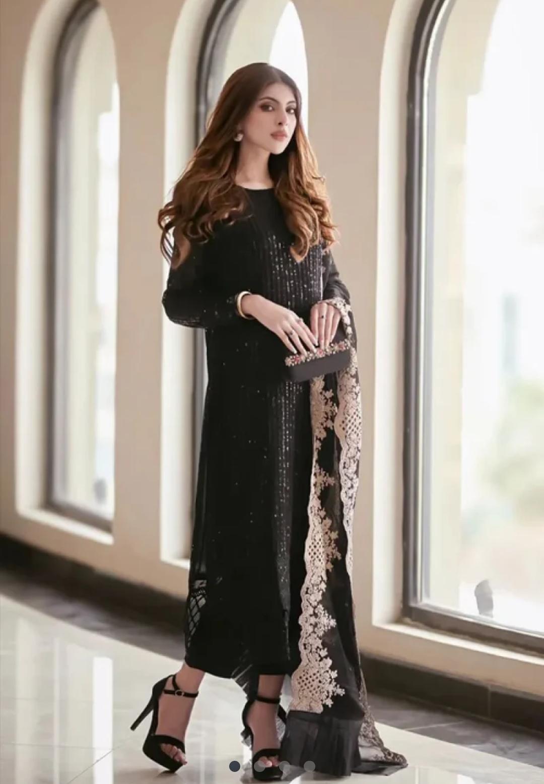 Woman in a black dress with lace details standing in a room with large windows.