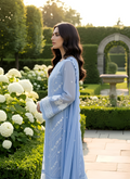 Woman in a light blue traditional outfit standing in a garden with white flowers and greenery.