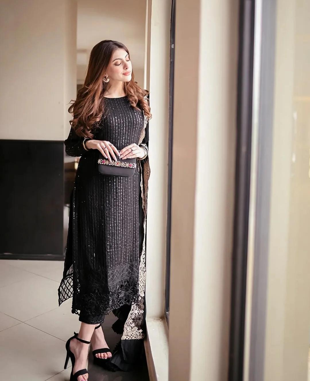 Woman in a black dress with lace details standing in a room with large windows.
