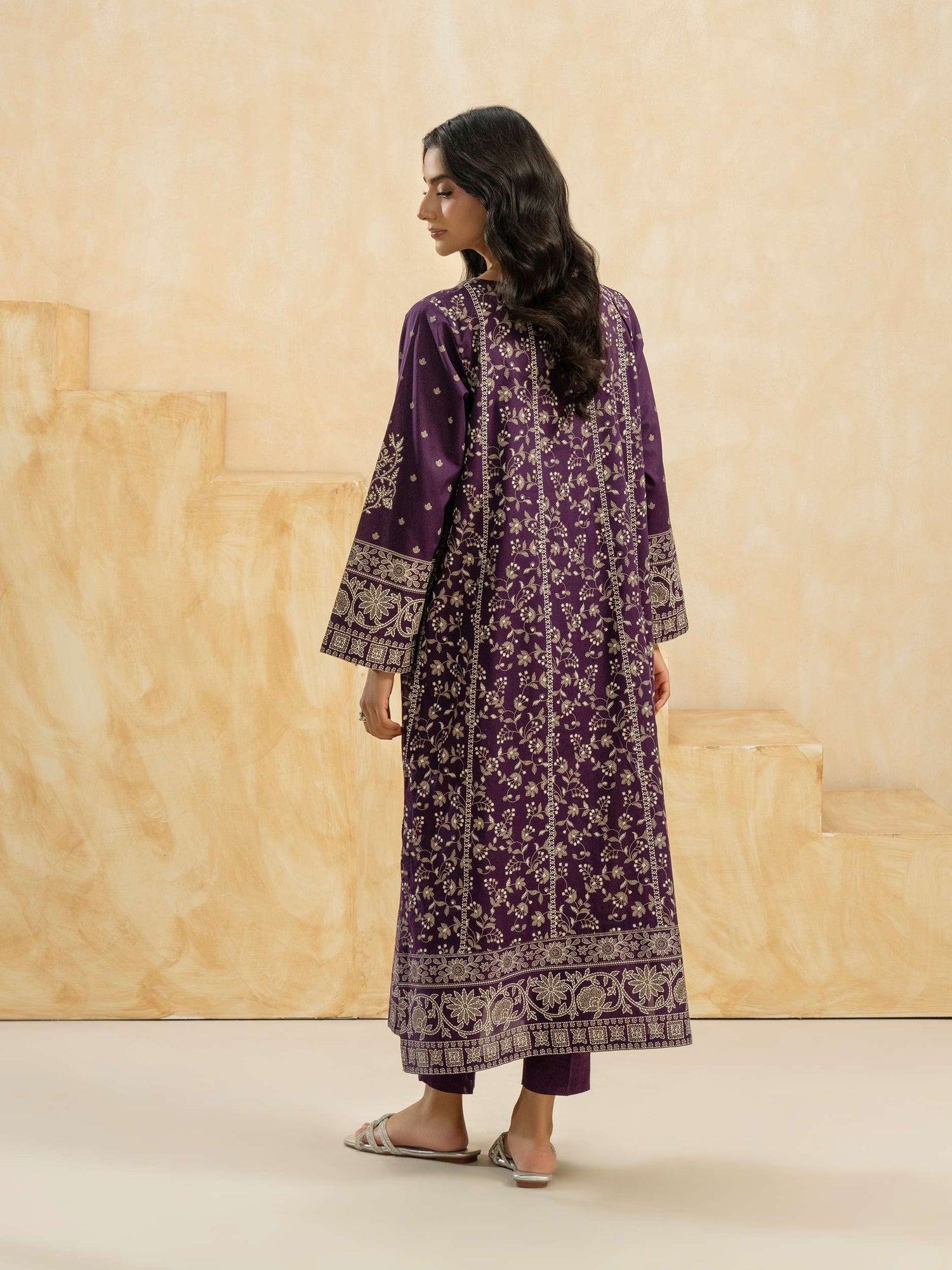Woman wearing a purple embroidered traditional outfit against a beige wall.