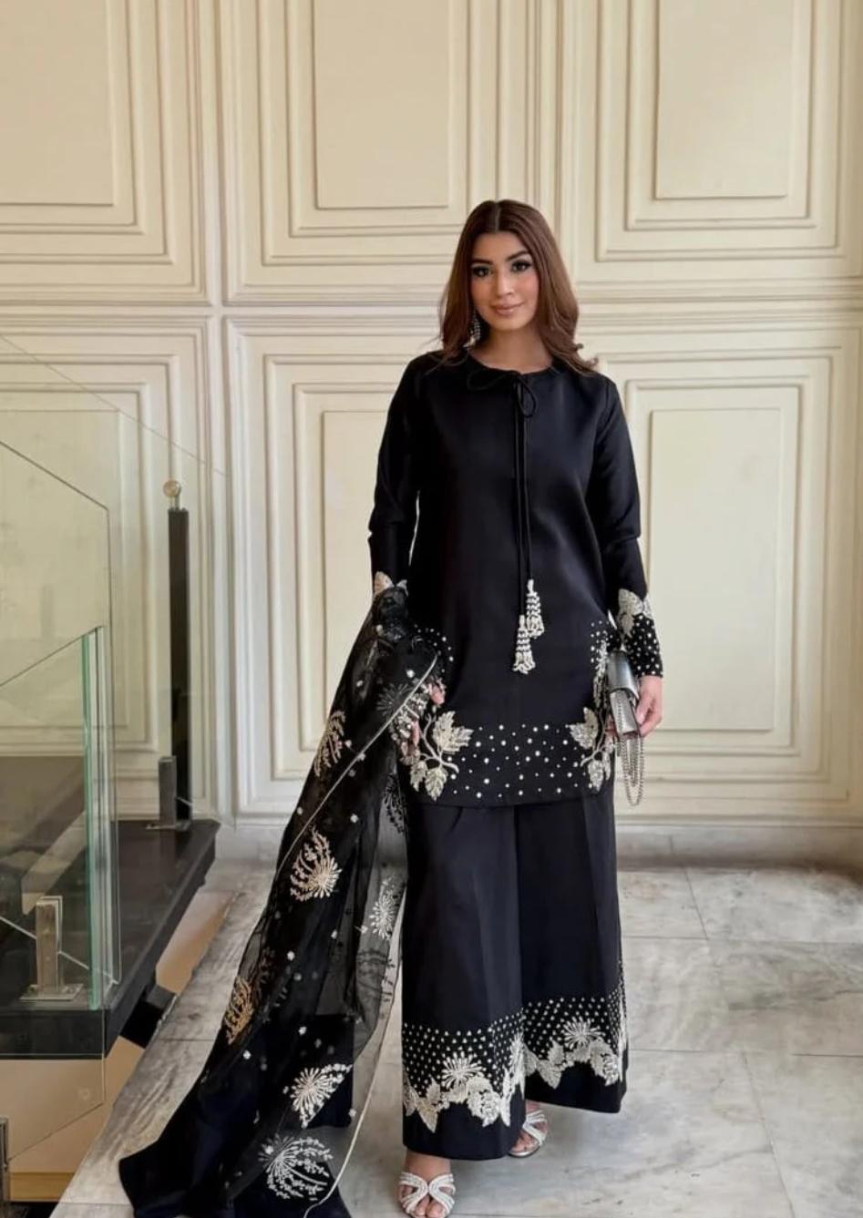 Woman in a black outfit with floral patterns standing in an elegant interior setting.