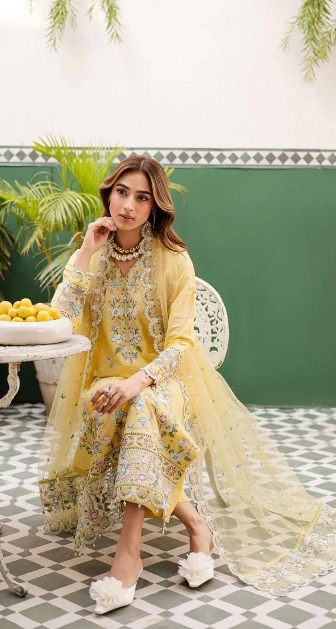 Woman in a yellow and white floral dress sitting on a chair with a green wall and plants in the background.